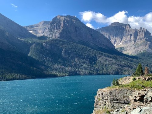 A serene lake surrounded by mountains under a blue sky with scattered clouds. The water is a vibrant turquoise.