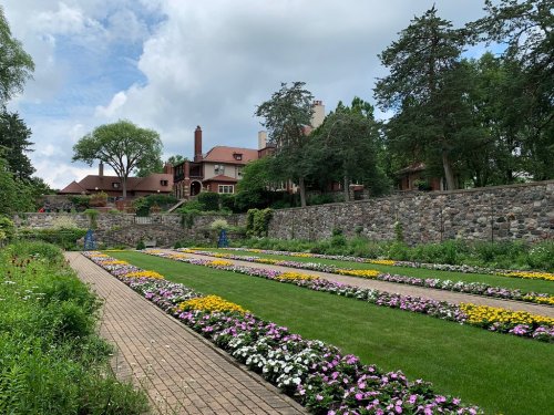 A landscaped garden with colorful flower beds and a stone wall, featuring a large house in the background under a cloudy sky.