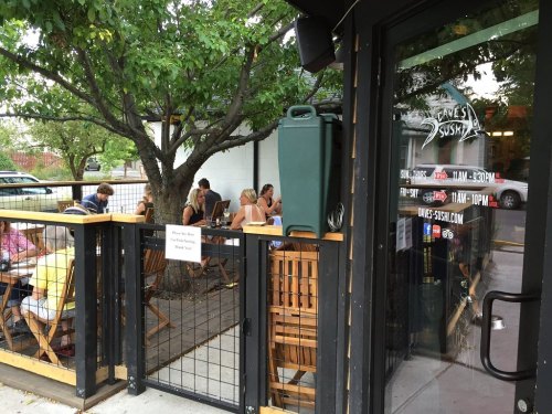 Outdoor dining area of a sushi restaurant with patrons seated at wooden tables under trees.