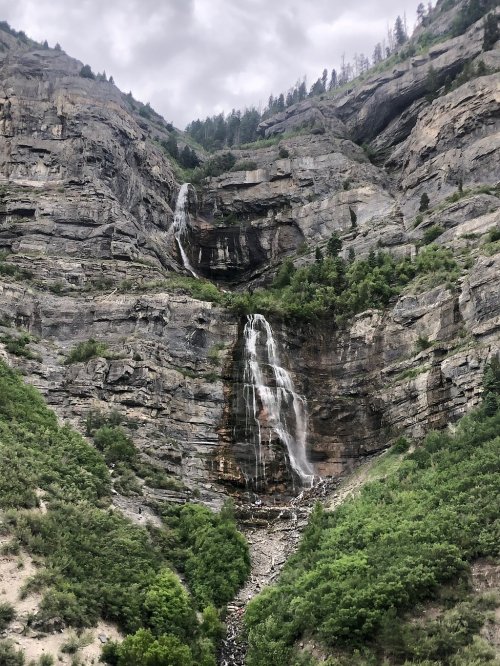 A majestic waterfall cascading down rocky cliffs, surrounded by lush greenery and a cloudy sky.