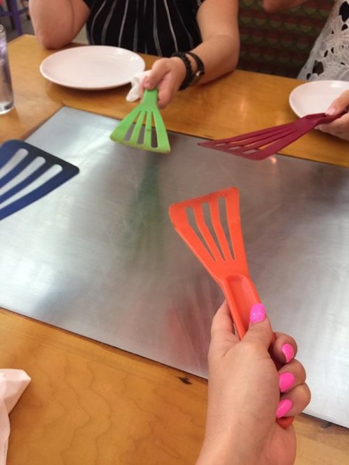 Hands holding colorful spatulas over a table with plates and a shiny surface, ready for cooking or serving.