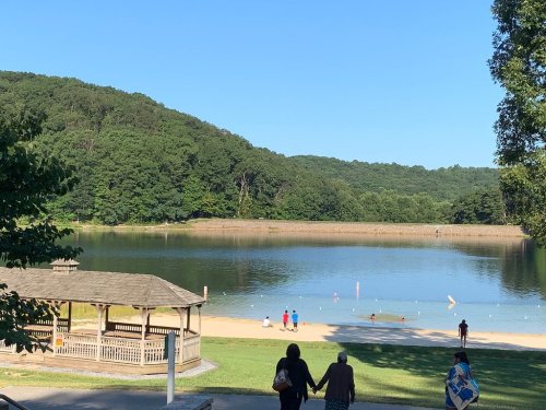 A serene lake with a sandy beach, surrounded by green hills, and people enjoying the water on a sunny day.