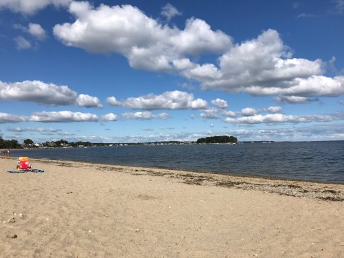 A sandy beach with calm water, scattered clouds, and a distant island under a bright blue sky.