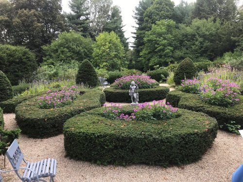 A lush garden with spiral hedges and vibrant flowers, featuring a statue in the center and surrounded by greenery.