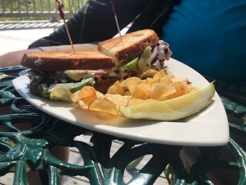A plate with a sandwich filled with meat and veggies, served with chips and a pickle slice, on a green table.