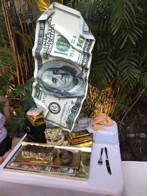 A lavish dining setup featuring a large dollar bill decoration, gold accents, and gourmet food on a table surrounded by greenery.