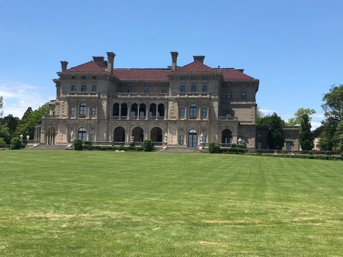 A grand mansion with a red-tiled roof, large balconies, and manicured lawns under a clear blue sky.