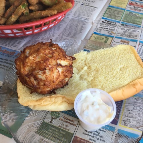 A fried crab cake on a bun with a side of mayonnaise, served on a clear plate over newspaper.