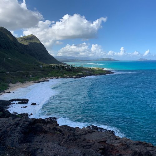 A scenic view of a coastline with turquoise waters, sandy beach, and lush green mountains under a partly cloudy sky.