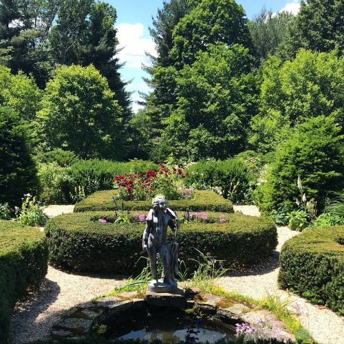 A serene garden with neatly trimmed hedges, colorful flowers, and a statue beside a small pond under a clear blue sky.