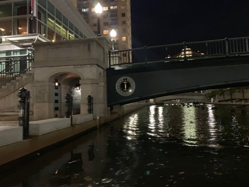 A nighttime view of a canal with a bridge, reflecting lights from nearby buildings and street lamps.