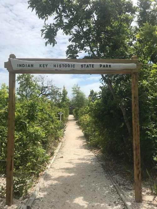 Sign for Indian Key Historic State Park at the entrance of a sandy path surrounded by lush greenery.