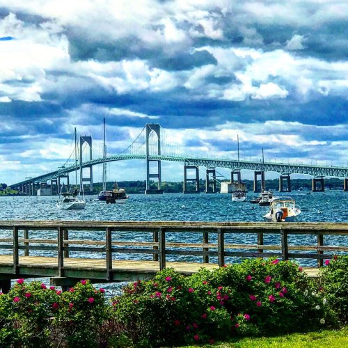 A scenic view of a bridge over water, with boats and a flower-filled foreground under a cloudy sky.