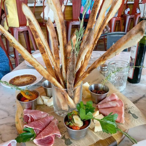 A platter featuring crispy breadsticks, cured meats, cheeses, and garnishes, served in a glass and on a wooden board.