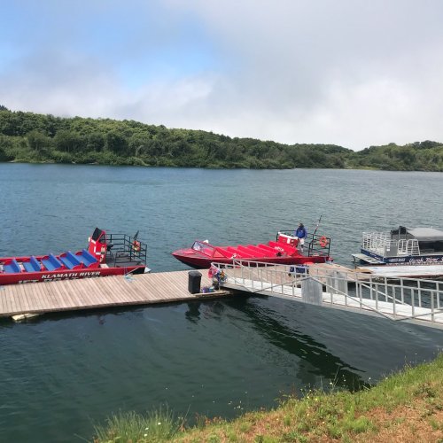 A dock with red and blue boats on a calm lake, surrounded by green trees and a cloudy sky.