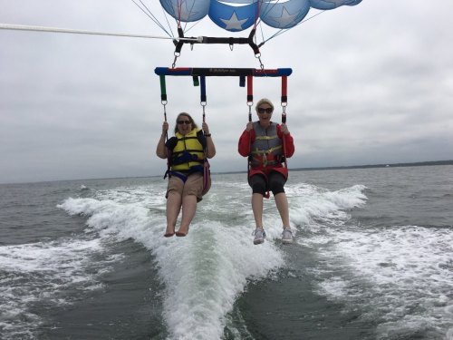 Two women parasailing over water, wearing life vests, with a cloudy sky in the background.