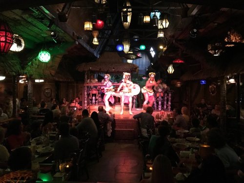 A lively performance with dancers in traditional attire on stage, surrounded by a vibrant, tropical-themed restaurant.