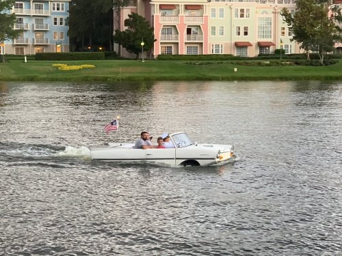 A couple enjoys a ride in a vintage car-boat on a calm lake, with colorful buildings in the background.