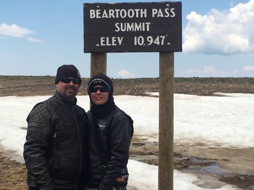 A couple in leather jackets stands by a sign for Beartooth Pass Summit, elevation 10,947, with snow in the background.