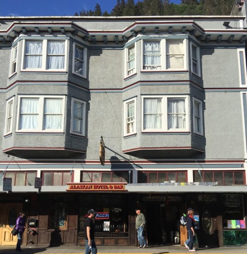 Historic building with large windows, featuring a sign for "Alaskan Hotel & Bar" and people walking by.