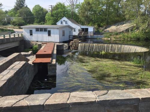 A small white building beside a river with a stone dam and lush greenery surrounding the water.