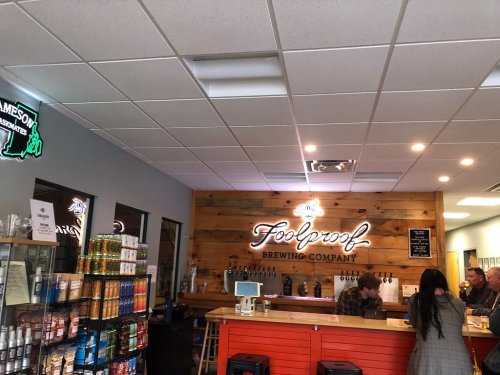 Interior of Foolproof Brewing Company, featuring a wooden bar, shelves of snacks, and patrons enjoying drinks.