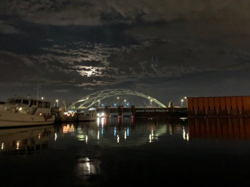 A nighttime view of a bridge over water, with boats docked and a full moon illuminating the cloudy sky.