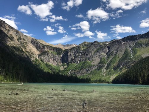 A serene lake surrounded by towering mountains and lush greenery under a blue sky with scattered clouds.