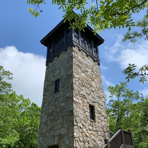 A tall stone tower with a wooden roof, surrounded by green trees and a blue sky with clouds.