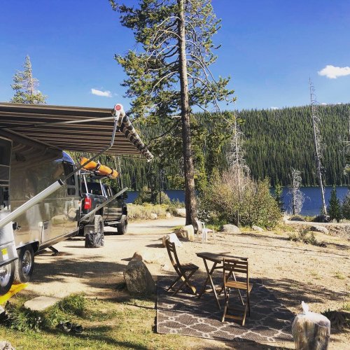 A scenic campsite with a silver trailer, wooden chairs, and a lake surrounded by trees under a clear blue sky.