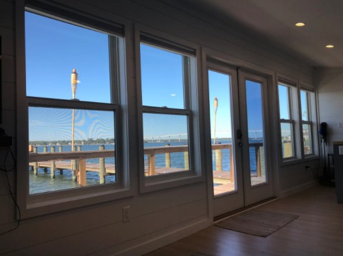 View of a serene waterfront through large windows, showcasing a wooden pier and clear blue sky.
