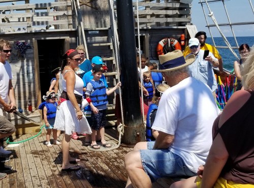 A group of children and adults on a pirate ship, some wearing life jackets, enjoying a sunny day at sea.