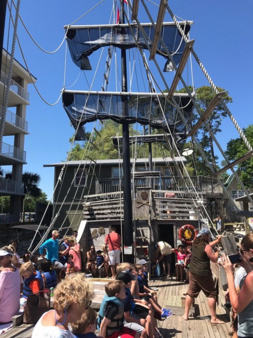 A pirate ship docked with children and adults gathered on the deck, enjoying a sunny day.