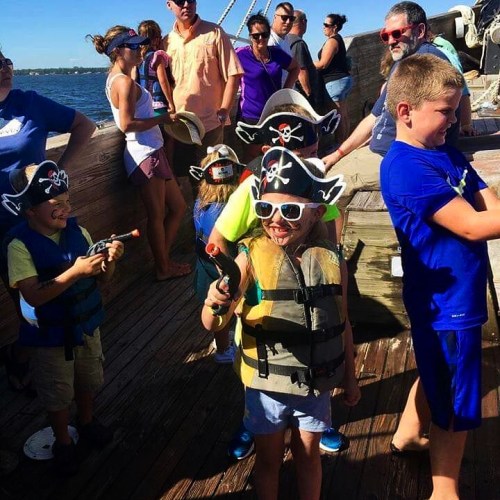 Children in pirate hats and life vests play on a ship, holding toy guns, with adults and a scenic water background.