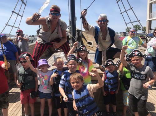A group of children in pirate hats and life vests pose with two pirate performers on a ship, smiling and cheering.