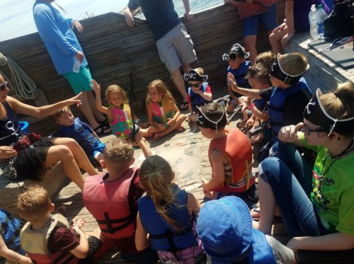 A group of children in life jackets gather around a map on a boat, listening to an adult leading an activity.