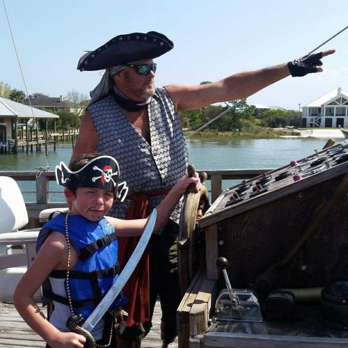 A pirate and a child in costume pose on a ship, with the pirate pointing towards the water.