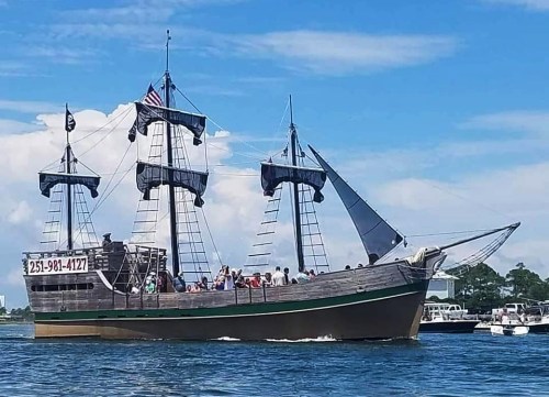 A large wooden pirate ship sails on blue water, with people on deck and a clear sky in the background.