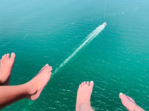 Two pairs of feet dangling above turquoise water, with a boat leaving a wake in the distance.