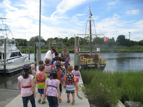 A group of children in pirate hats walks toward a docked pirate ship by a river, with a guide in front.