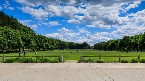 A spacious park with green grass, trees, and people enjoying the outdoors under a blue sky with fluffy clouds.