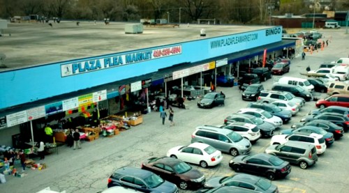 Aerial view of Plaza Flea Market with shoppers and parked cars in a busy lot. Bright blue signage and various stalls visible.