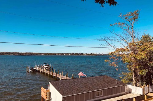 A serene lake view with a dock, a boat, and a clear blue sky, framed by trees and a nearby house.