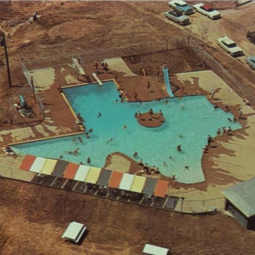 Aerial view of a swimming pool area with people swimming, a slide, and surrounding vehicles.