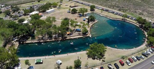 Aerial view of a park with a winding river, people swimming, and cars parked along the road. Green trees surround the area.