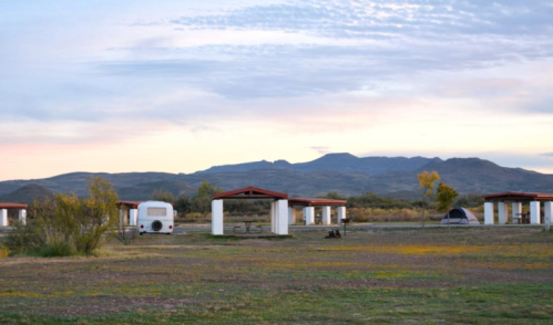 A scenic view of a campsite with white structures, a trailer, and mountains in the background under a colorful sky.