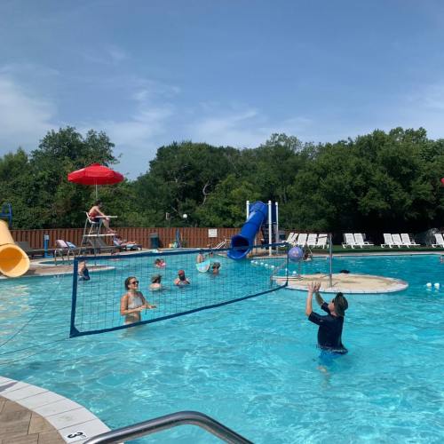 A sunny pool scene with people swimming, playing volleyball, and enjoying water slides under a clear blue sky.