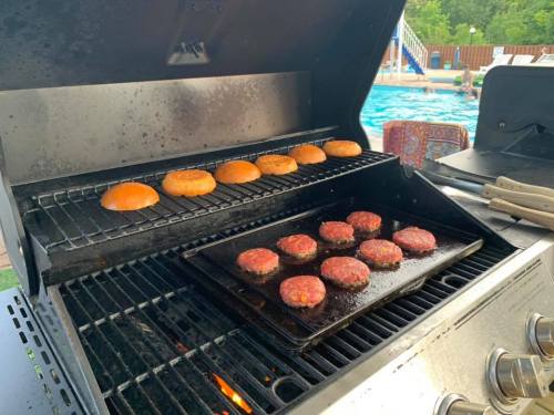 Grill with burgers and buns cooking, with a pool and lounge chairs visible in the background.