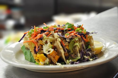 Three colorful tacos filled with vegetables, served on a plate with a small cup of sauce.