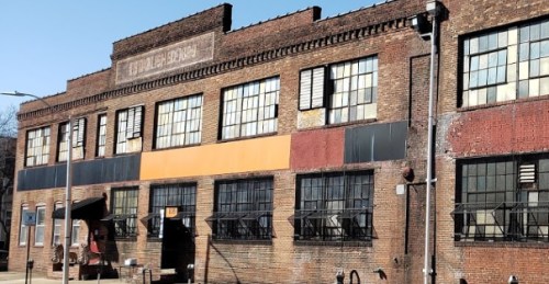 A brick building with large windows, some boarded up, featuring a faded sign and industrial elements.